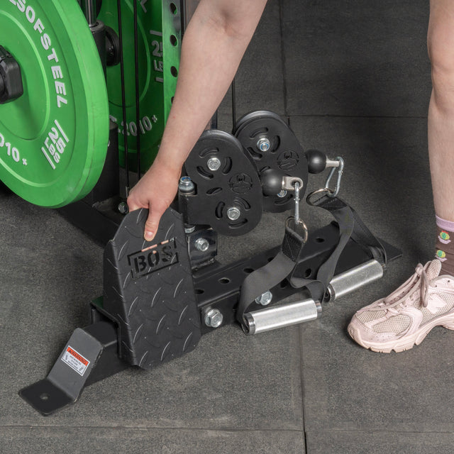 A person in pink sneakers and flower socks adjusts a Bells of Steel Footplate for Cable Tower/All In One Trainer on a gym machine, with green weight plates close by.