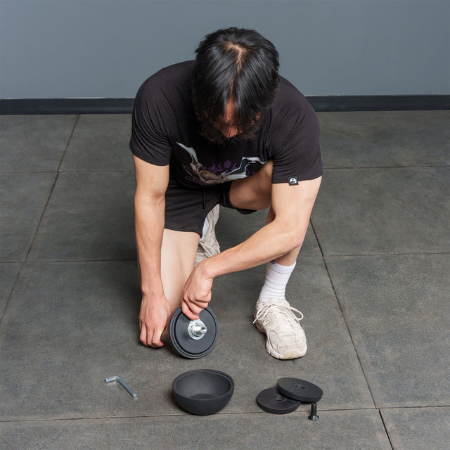 A person kneels on a tiled floor assembling the Bells of Steel 6-12KG Adjustable Kettlebell, with plates, a wrench, and a screw-on collar laid out nearbyâa detailed setup reminiscent of demonstrations by Mark Wildman when showcasing adjustable kettlebell equipment.