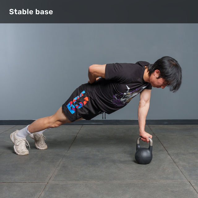 Wearing workout clothes, a person performs a one-arm plank on the Bells of Steel 6-12KG Adjustable Kettlebell, with their other hand behind their back, on a gray-tiled gym floor. Text at top reads âStable base.â.