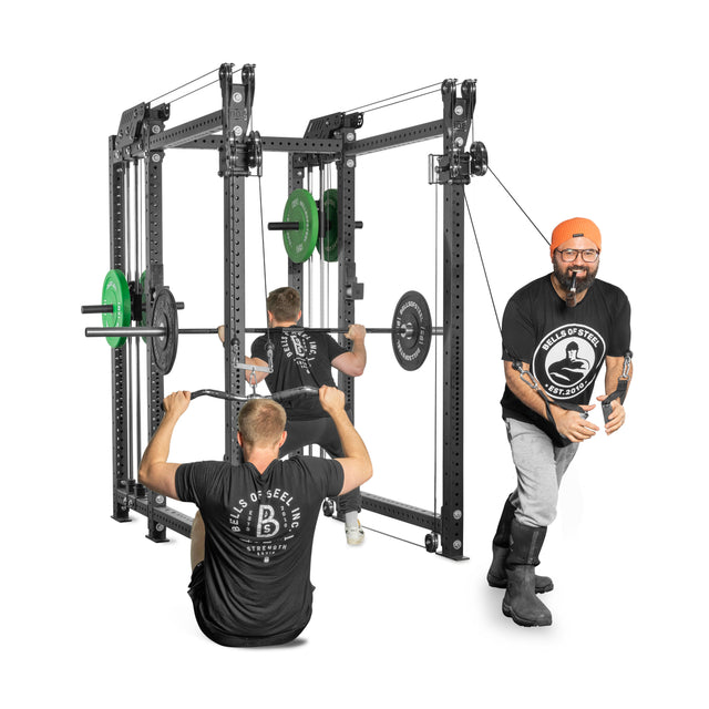 Two men work out using the Kraken Rack Attachment for 6 Post Manticore Racks (Single Side) by Bells of Steel. One, seated, uses the pulley system while the other stands by a dual-handle cable tower. Both wear black shirts with white logos. A third person in an orange hat smiles in the foreground.