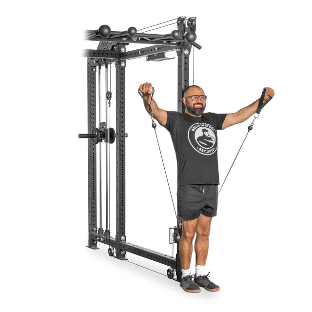 A person with glasses is working out on the Kraken Rack Attachment for 6 Post Manticore Racks from Bells of Steel. They're in a black t-shirt, shorts, and sneakers, pulling cables outward while the compact system's weights and adjustable trolleys optimize their exercise.