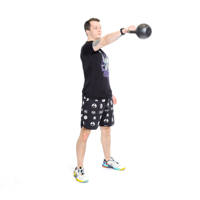 A man stands on a white background, holding a kettlebell at shoulder height with one arm forward. He wears Bells of Steel Gym Shorts, a black T-shirt, and colorful sneakers designed for comfort and agility.