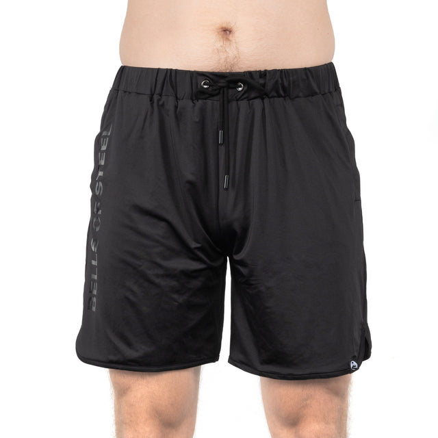 A person wears Bells of Steel Gym Shorts with a drawstring waist and stretchy fabric. "BELLS OF STEEL" is printed vertically on the left leg. The photo is cropped from mid-torso to above the knees.