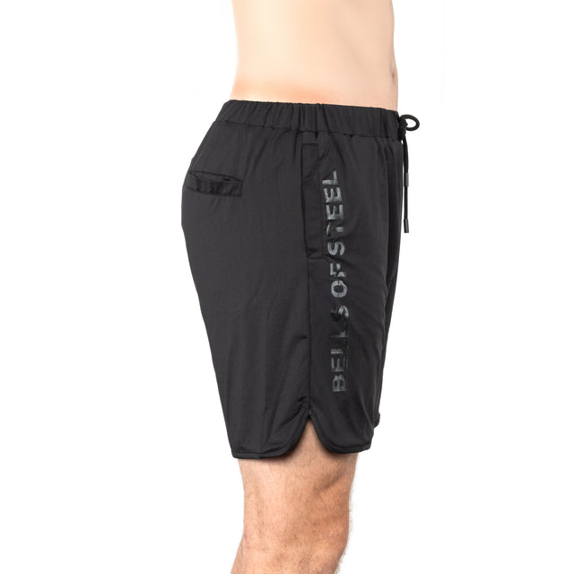 A person in Bells of Steel Gym Shorts, featuring stretchy black fabric and vertical "BELLS OF STEEL" print on the left side, is shown in a side profile against a white background.