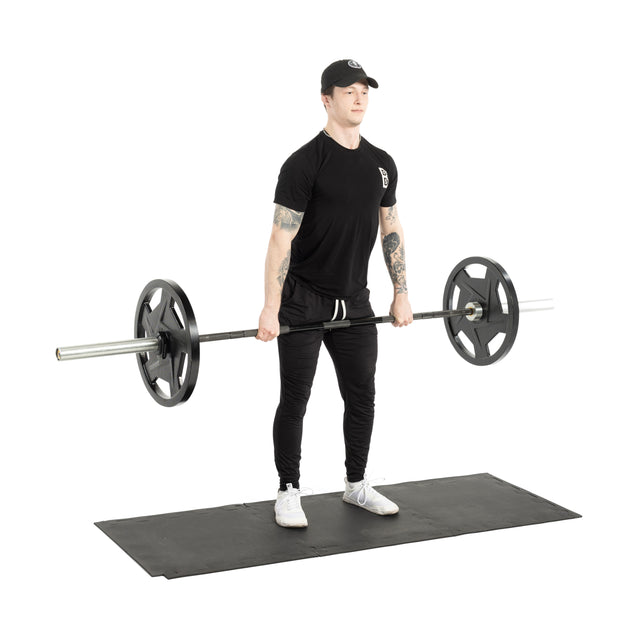 A person stands on Bells of Steel 24" x 24" Puzzle Mats (Set of 8), wearing black attire and holding a barbell loaded with weights, getting ready to perform a deadlift.