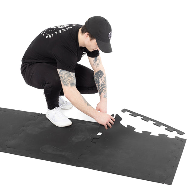 A person in black clothing and a cap is kneeling on a white background, assembling edge pieces of Bells of Steel 24" x 24" Puzzle Mats (Set of 8) for gym floor protection.