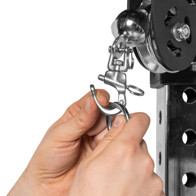 Hands are securing a Bells of Steel Swivel Shackles carabiner clip onto a metal hook attached to a pulley system on a perforated metallic structure.