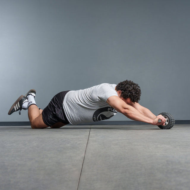 A man with curly hair kneels on a gym floor, wearing a gray t-shirt and black shorts, using the Bells of Steel ABsyidyan Stainless Steel Ab Roller with an extra-wide dual wheel to extend his body forward.