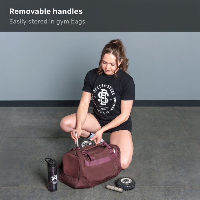A woman kneels, packing the Bells of Steel ABsyidyan Stainless Steel Ab Roller with extra-wide dual wheels into a maroon gym bag. Nearby are a water bottle and two wheels. Text: "Removable handles—easily stored in gym bags.