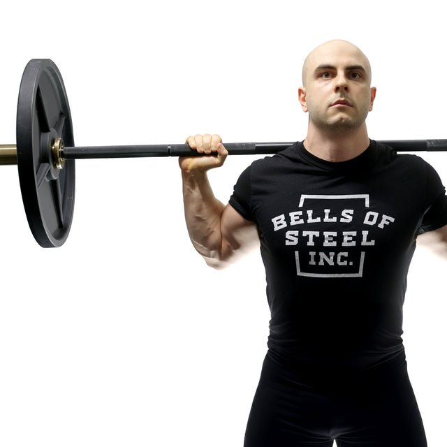 A muscular, bald man in a Bells of Steel t-shirt holds the Alex Leonidas Onyx Bar with a Cerakote finish across his shoulders, preparing to squat against a plain white background.