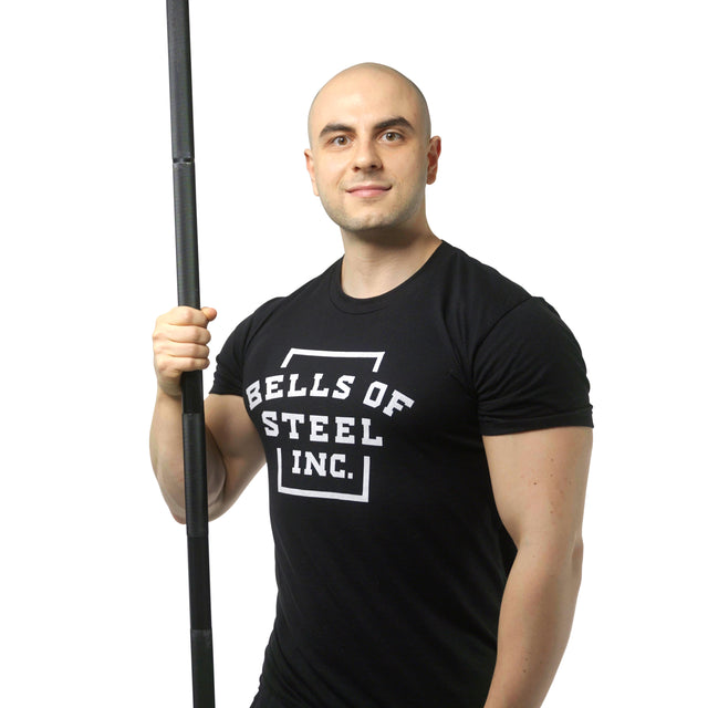 A bald man in a black Bells of Steel t-shirt stands against a white background, smiling slightly as he holds a vertically positioned Alex Leonidas Onyx Bar from Bells of Steel with a Cerakote finish.