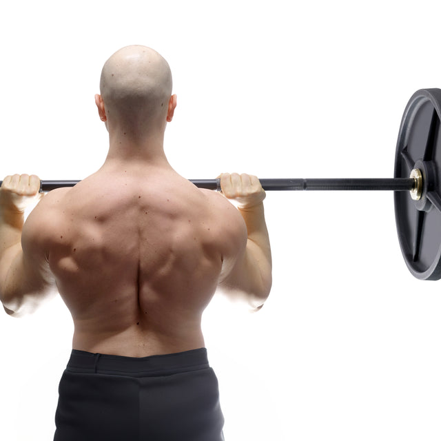 A muscular, bald man lifts a Bells of Steel Alex Leonidas Onyx Bar loaded with weights across his shoulders, displaying defined back and arm muscles against a white background.