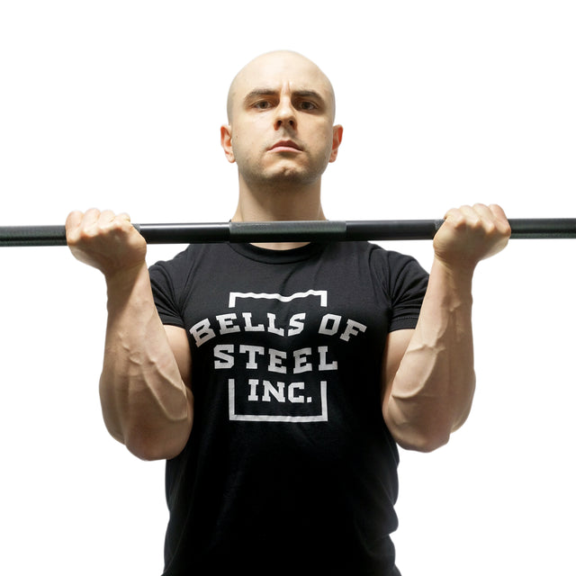 Wearing a Bells of Steel t-shirt, a person holds the Alex Leonidas Onyx Bar at chest level with bent elbows, set against a plain white background.