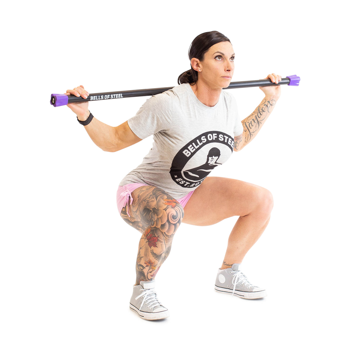 A person crouches with a Weighted Workout Body Bar across their shoulders, demonstrating their strength training skills. They're sporting a "Bells of Steel" t-shirt, pink shorts, and high-top sneakers as part of their fitness gear, with intricate tattoos gracing their right leg.