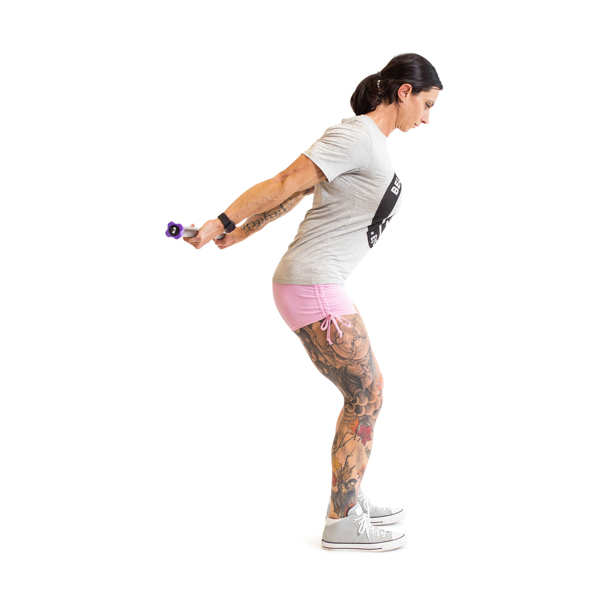 A tattooed individual, dressed in a gray t-shirt, pink shorts, and sneakers, engages in a strength training exercise using Weighted Workout Body Bars by Bells of Steel. They maintain a stance with knees slightly bent and arms extended backward, all set against a white background.