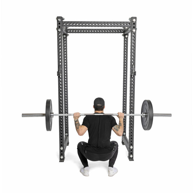 Wearing black athletic gear and a cap, a person squats with Bells of Steel All-Black Bumper Plates on a barbell inside a squat rack, seen from behind against a white background.