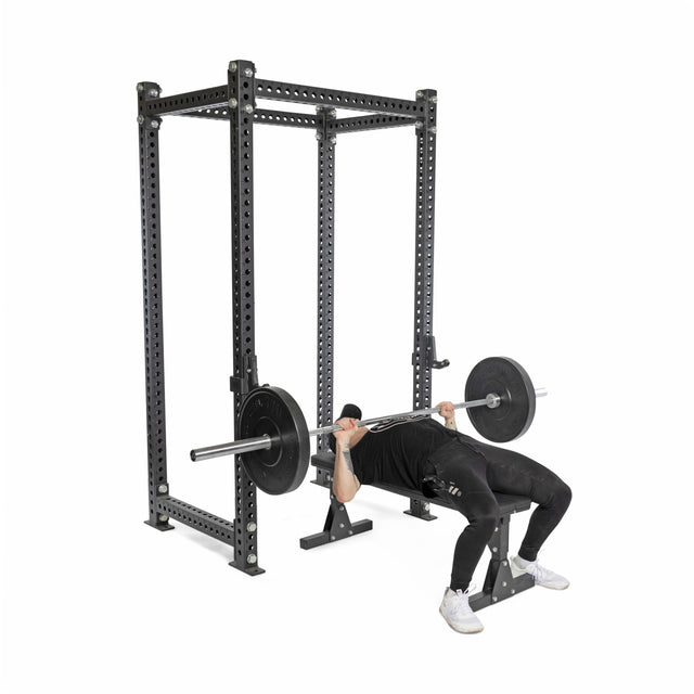 A person in black workout gear and white shoes is bench pressing with Bells of Steel All-Black Bumper Plate Sets on a bench inside a power rack against a plain white background, emphasizing lifting safety.