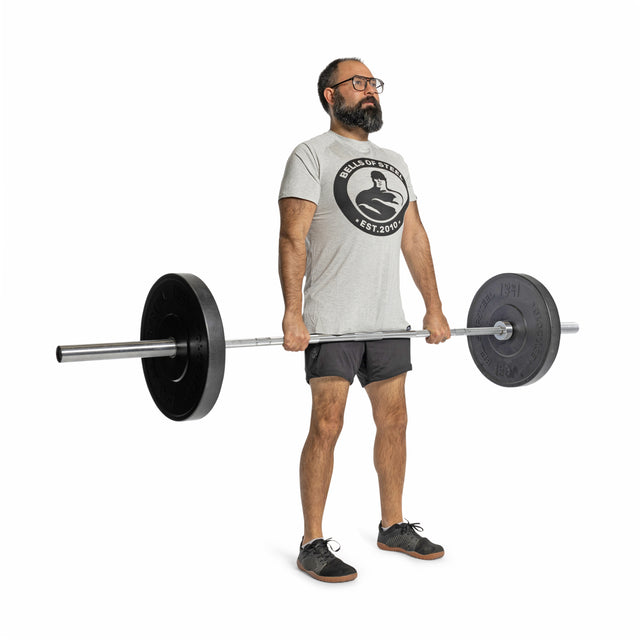 A bearded man with glasses wears a gray t-shirt, black shorts, and sneakers as he stands against a white background holding a barbell loaded with Bells of Steel All-Black Bumper Plates, preparing for a lift.