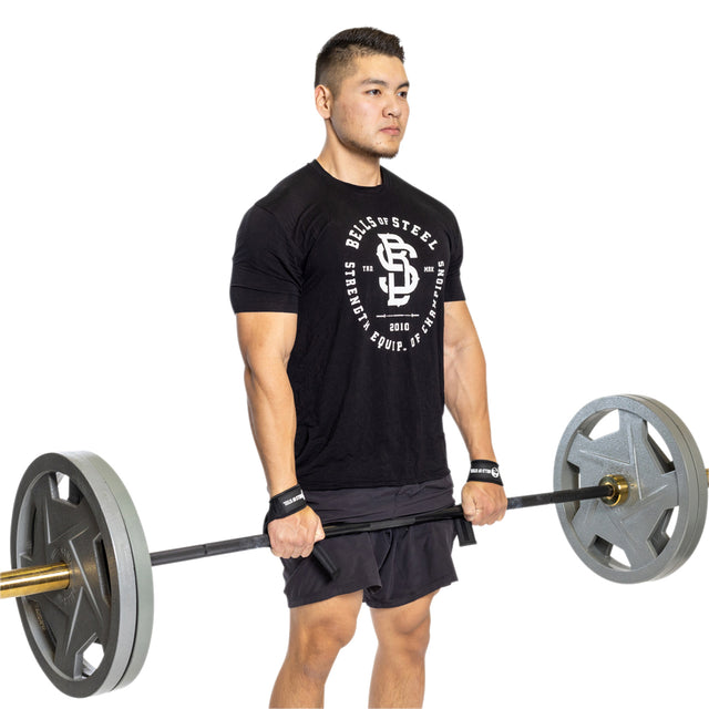 A man demonstrates grip strength using Bells of Steel Lifting Straps as he holds a loaded barbell at thigh level, wearing a black T-shirt and shorts on a white background.