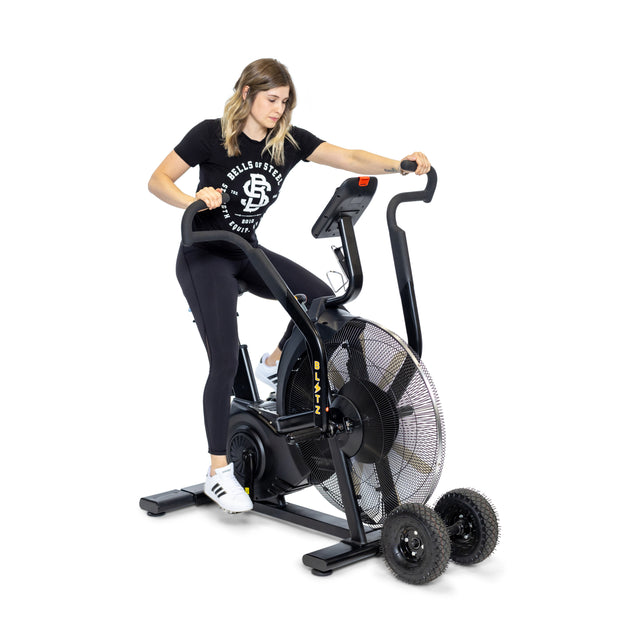 A woman in a black T-shirt and leggings sits on the Bells of Steel Blitz Max Air Bike, resting one hand on the handlebar. Two spare wheels are placed beside the bike, set against a white background.