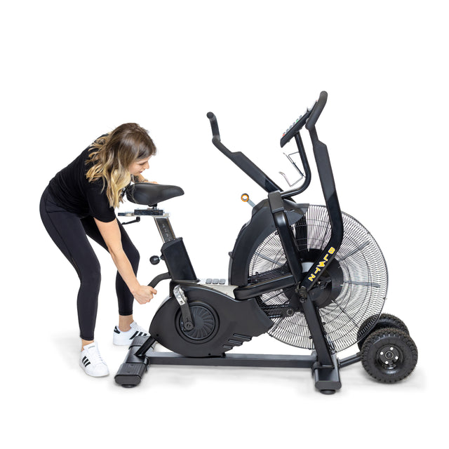 A woman in athletic wear adjusts the seat height on a Bells of Steel Blitz Max Air Bike with large fan wheels, set against a plain white background.