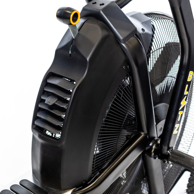 Close-up of the black Bells of Steel Blitz Max Air Bike fan with a protective cover and part of the pedal visible. This commercial air bike features bold yellow "BLITZ" branding, making it perfect for any fitness studio equipment lineup.