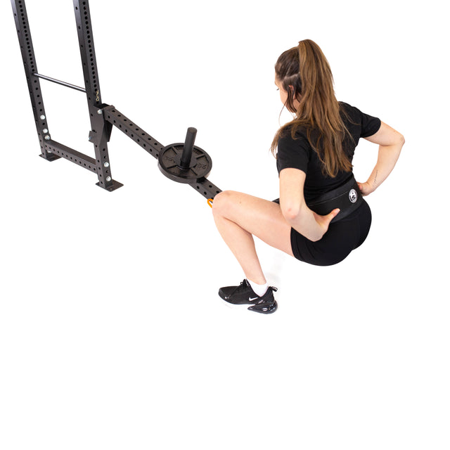 In bright lighting, someone in black athletic wear and headphones does strength exercises with the Bells of Steel Belt Squat/Lever Arms Rack Attachment on a power rack, crouching low while keeping one hand on their back.