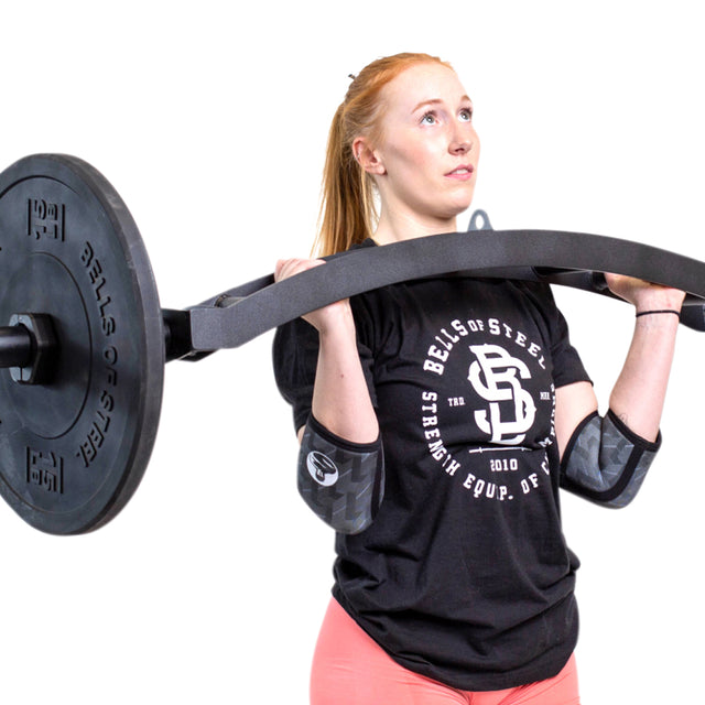 A red-haired woman in a black Bells of Steel shirt and pink leggings lifts a curved barbell to her shoulders while wearing Bells of Steel Elbow Sleeves for support, set against a white background.