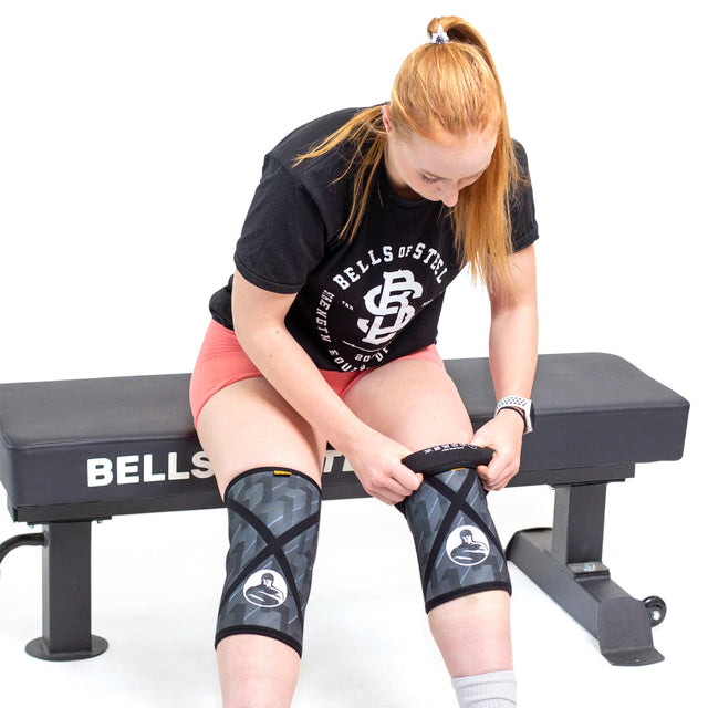 A woman with long red hair sits on a workout bench, wearing a black Bells of Steel t-shirt and pink shorts as she adjusts Bells of Steel Knee Sleeves for added squat support on both legs.