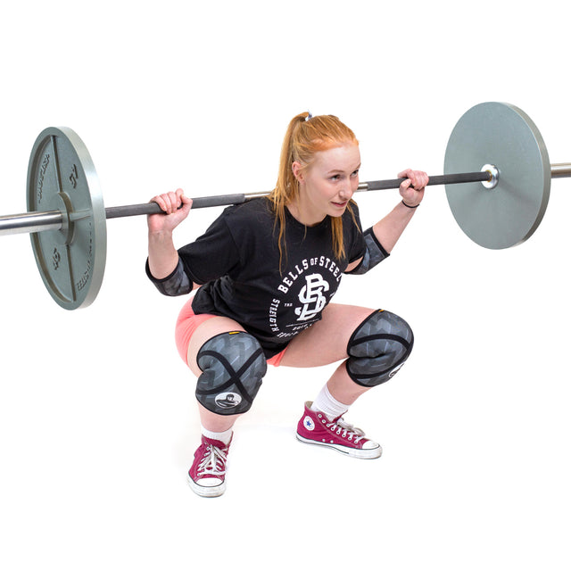 A woman with long red hair, wearing Bells of Steel Knee Sleeves, a black shirt, pink shorts, and red sneakers, squats while lifting a barbell with weights. She looks focused during her workout against a white background.