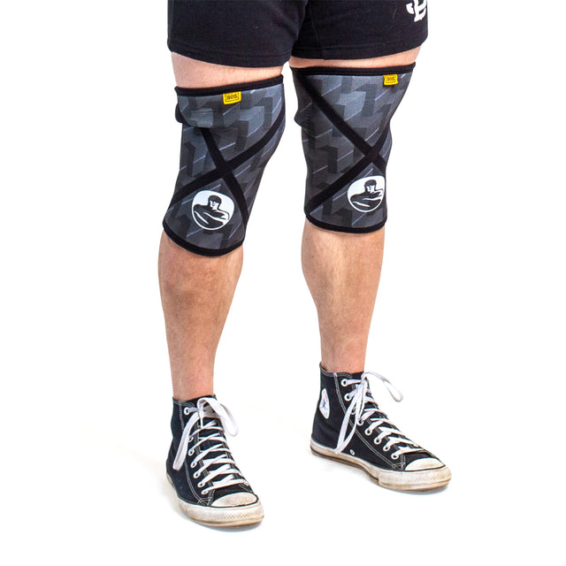 A person wears Bells of Steel Knee Sleeves with a geometric design, styled with black athletic shorts and black high-top sneakers with white laces, standing against a white background, ready for squat support.