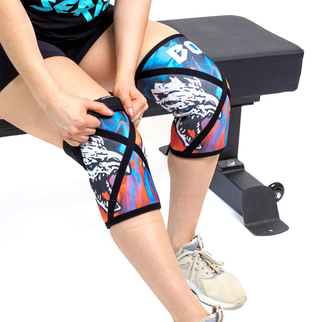 A person in athletic gear sits on a gym bench, adjusting Bells of Steel Knee Sleeves with a vibrant wolf design on each knee, paired with beige sneakers.