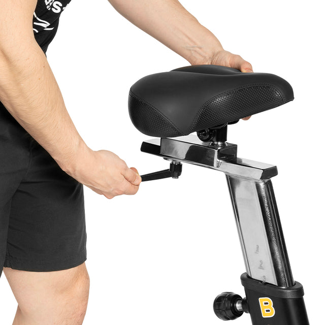 A user adjusts the seat height of the Bells of Steel Blitz Air Bike with a side knob. The bike seat and part of their arm and shorts are visible—ideal for customizing full-body home gym workouts.