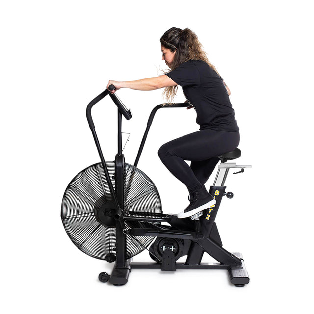 A woman in black athletic wear uses the Bells of Steel Blitz Air Bike, gripping the handles and pedaling for a full-body workout against a white background—ideal for any home gym.