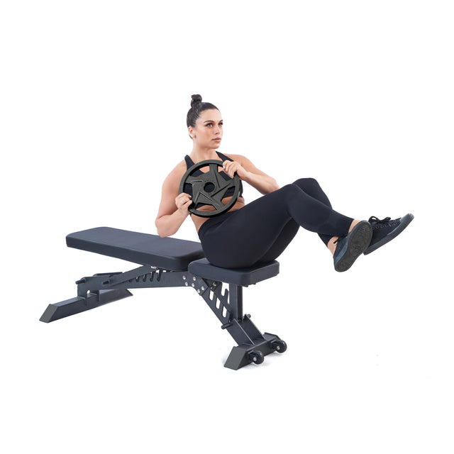 Clad in black attire, a person performs a Russian twist on a workout bench, showcasing core strength. Holding Black Mighty Grip Olympic Weight Plates from Bells of Steel, they demonstrate dedication to form and strength balance with lifted legs.