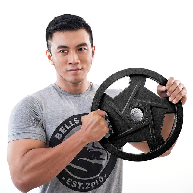 A person in a gray T-shirt holds a Bells of Steel Black Mighty Grip Olympic Weight Plate with precision grip handles, visible ridges, and text against a plain white background.