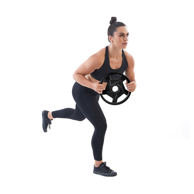 A woman in a black workout outfit performs a balance exercise with Bells of Steel Black Mighty Grip Olympic Weight Plates, holding them with precision grip handles. She stands on one leg, her other leg bent behind, set against a plain white background.