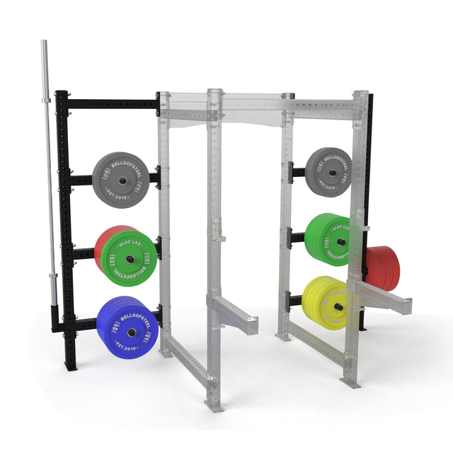 The Bells of Steel Hydra Batwing Storage squat rack features sturdy 3x3 uprights and a black and silver frame, loaded with gray, blue, green, yellow, and red weight plates. A barbell is mounted on the left side of the rack.