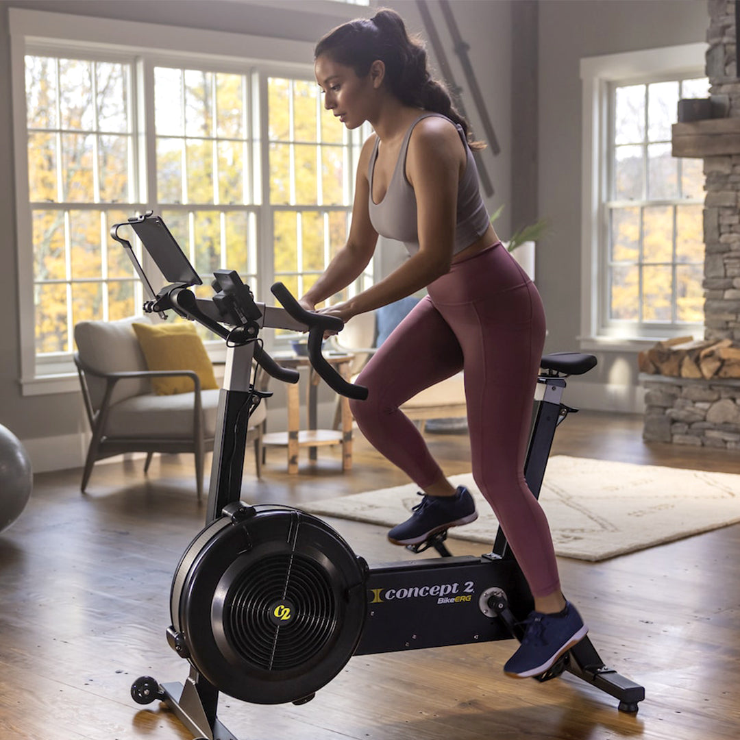 A woman enjoys a full-body, low-impact workout on a Concept 2 BikeERG with PM5 in her sunlit living room. Dressed in a light tank top and pink leggings, she pedals with focus. A large window frames the autumn trees outside, while a yoga ball and armchair sit quietly in the background.