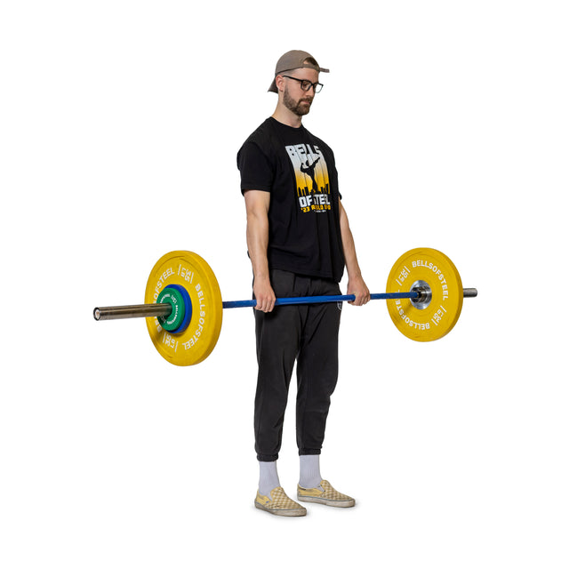 A man in a cap, glasses, black T-shirt and pants, and plaid shoes faces forward holding a barbell with yellow Bells of Steel Urethane-Coated Change Plates on each end, set against a plain white background.