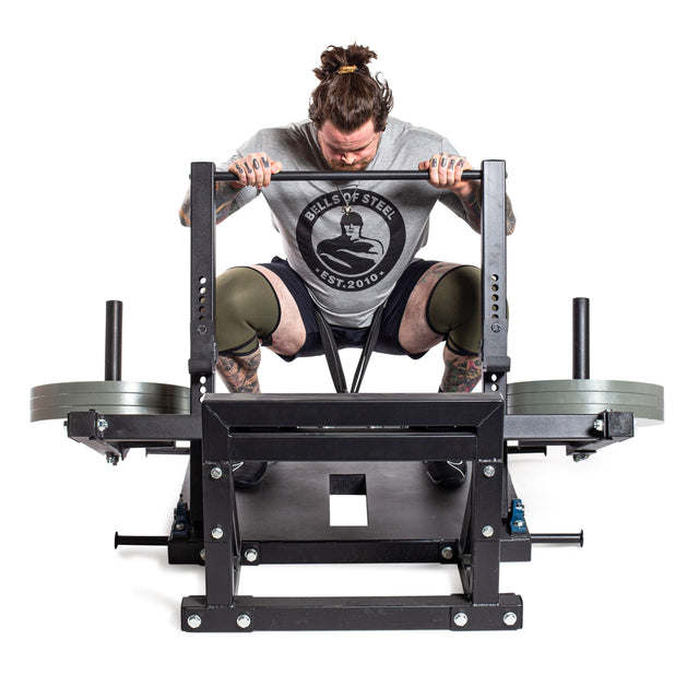 Wearing knee sleeves and a "Bells of Steel" shirt, a tattooed lifter grips the handles of a belt squat machine loaded with Bells of Steel Machined Iron Olympic Weight Plate Sets, focusing intensely before their lift.