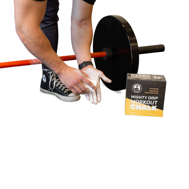 A person applies Bells of Steel Mighty Grip Workout Chalk to their hands beside a barbell with black weight plates, while the product box rests on the floor.