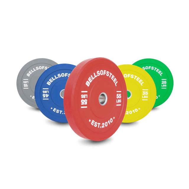 Five Bells of Steel Coloured Bumper Plate Sets (15, 25, 35, 45, and 55 lbs; gray, blue, red, yellow, green) are arranged upright in a row. Each plate features a durable stainless steel insert ring.