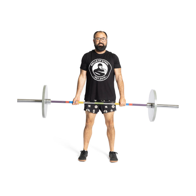 A bearded man with glasses, in a black "Cups of Steel" shirt, patterned shorts, and black shoes, stands holding the Bells of Steel Hydra Seven Ring Bar with weights on each end against a white background.