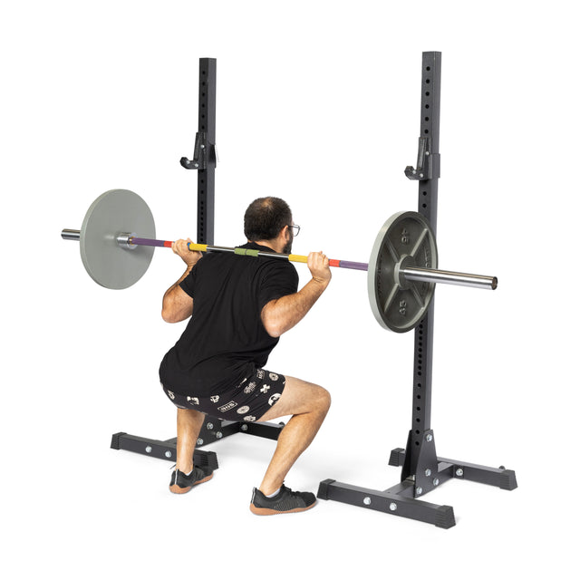 A person in a black shirt and patterned shorts performs a barbell squat on a squat rack, gripping the Bells of Steel Hydra Seven Ring Bar with hands on its placement rings and keeping their heels flat.