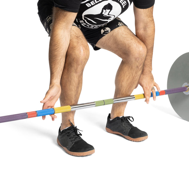 A person in black sneakers and shorts prepares to lift the Bells of Steel Hydra Seven Ring Bar, which has colorful grips and visible hand placement rings, viewed from the waist down against a white background.