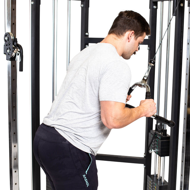 A man in a light gray t-shirt and black shorts performs a triceps pushdown using the Bells of Steel Multi Purpose Close Grip Cable Attachment, gripping the bar with both hands on a cable machine.