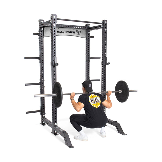 A person in a black shirt and cap performs a barbell back squat inside the Bells of Steel Manticore Collegiate Power Rack Builder (3" x 3", 1" Holes) in their home gym, viewed from behind.