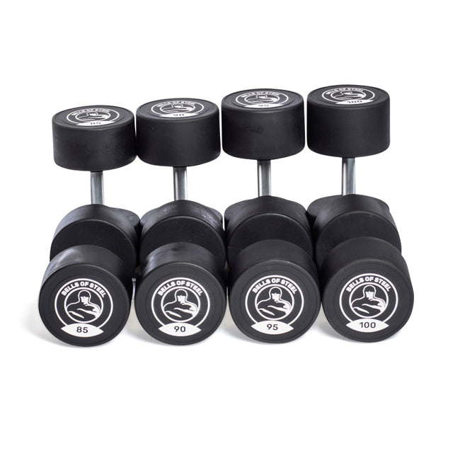 Five pairs of Bells of Steel Urethane Dumbbells are arranged in a row with weights 85, 90, 95, and 100 lbs. Each has circular ends featuring a logo that highlights their commercial-grade quality.
