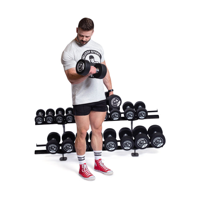 A man in a gray T-shirt and black shorts lifts a Bells of Steel Urethane Dumbbell with his right arm. He's wearing red shoes and stands before a rack lined with urethane dumbbells against a plain white background.